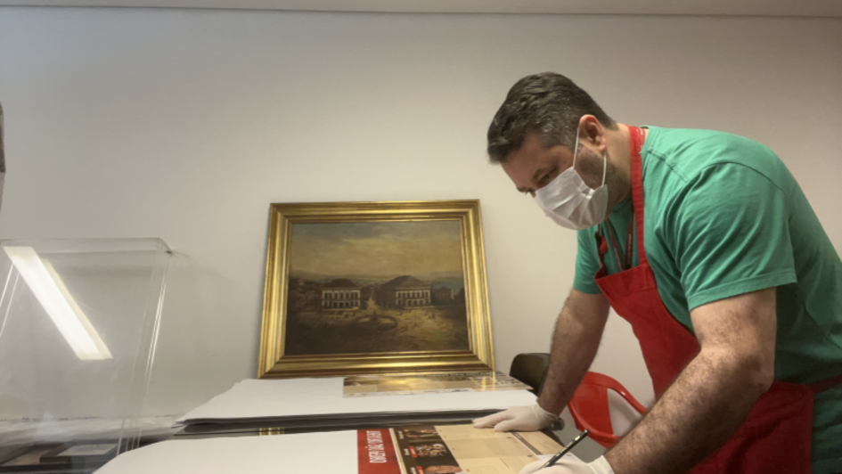 Homem usando m&aacute;scara cir&uacute;rgica, luvas brancas e avental vermelho trabalha sobre uma mesa, preenchendo ou assinando um material impresso de grande formato. Ao fundo, h&aacute; um quadro emoldurado apoiado na parede e uma cadeira vermelha. O ambiente parece ser um espa&ccedil;o de conserva&ccedil;&atilde;o, cataloga&ccedil;&atilde;o ou prepara&ccedil;&atilde;o de documentos e obras.