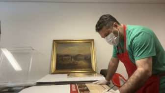 Homem usando m&aacute;scara cir&uacute;rgica, luvas brancas e avental vermelho trabalha sobre uma mesa, preenchendo ou assinando um material impresso de grande formato. Ao fundo, h&aacute; um quadro emoldurado apoiado na parede e uma cadeira vermelha. O ambiente parece ser um espa&ccedil;o de conserva&ccedil;&atilde;o, cataloga&ccedil;&atilde;o ou prepara&ccedil;&atilde;o de documentos e obras.